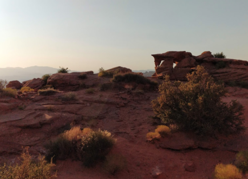 A rocky landscape at sunset, featuring red rock formations and sparse vegetation under a clear sky.