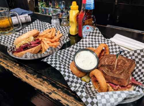 A table with two plates: one with a sandwich and onion rings, the other with fries and a hot dog, plus drinks.
