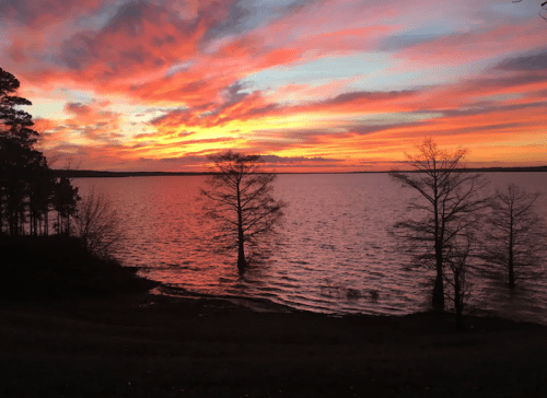 A vibrant sunset over a lake, with colorful clouds and silhouetted trees along the shoreline.