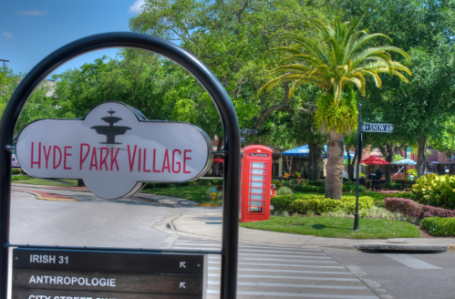 Sign for Hyde Park Village with palm trees and a red telephone booth in the background on a sunny day.