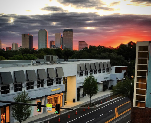 City skyline at sunset with clouds, featuring buildings and a street lined with trees and traffic cones.
