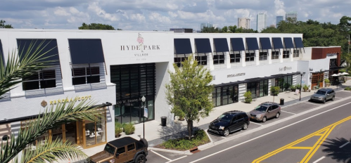 Aerial view of Hyde Park Village, featuring shops and greenery, with a city skyline in the background.