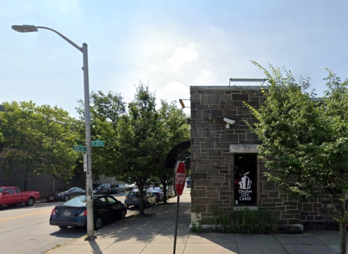 A stone building with a cake shop sign, surrounded by trees and parked cars on a sunny day.
