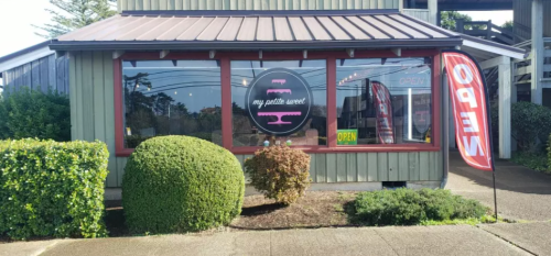 Exterior of a bakery with a round sign reading "my petite sweet" and an "OPEN" flag outside. Green shrubs in front.
