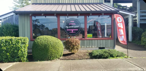 Exterior of a shop named "my petite sweet," featuring an open sign and a colorful flag. Lush greenery surrounds the entrance.
