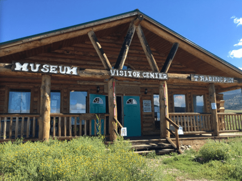 Log cabin-style building with signs for a museum, visitor center, and trading post, surrounded by greenery and blue sky.