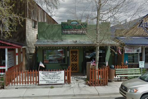 A green storefront with a sign reading "Among Friends" and a wooden fence, featuring flags and a bench outside.