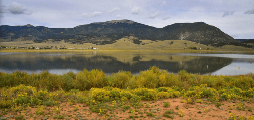 A serene lake reflects mountains and clouds, surrounded by lush greenery and wildflowers in a tranquil landscape.