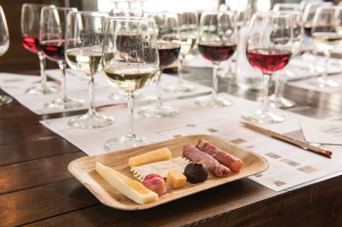 A wooden platter with assorted cheeses and meats, surrounded by wine glasses filled with red and white wine.