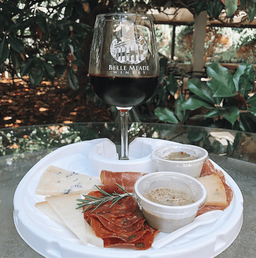 A glass of red wine from Belle Meade Winery beside a plate of assorted cheeses, meats, and dips on a table.