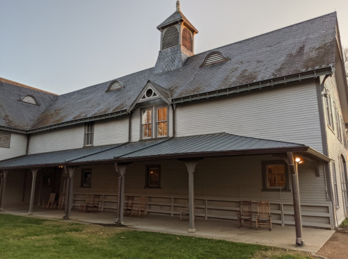 A historic building with a peaked roof, porch, and rocking chairs, set against a twilight sky.