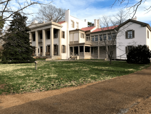 Historic mansion with a mix of architectural styles, surrounded by green grass and trees under a blue sky.