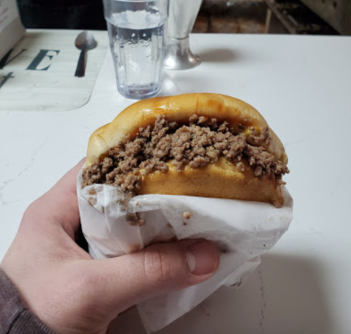 A hand holding a sandwich filled with ground beef, wrapped in paper, with a glass of water in the background.