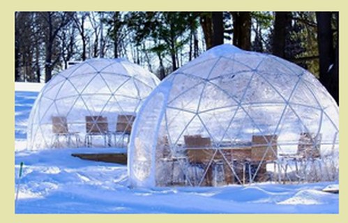 Two transparent geodesic domes in a snowy landscape, each with wooden tables and chairs inside.