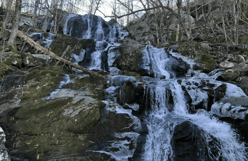 A cascading waterfall flows over rocky terrain, surrounded by bare trees and a rugged landscape.