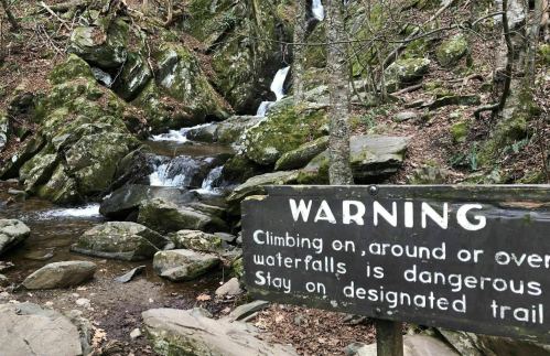 A warning sign near a rocky area with a small waterfall, advising caution and to stay on designated trails.