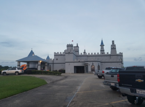 A gray castle-like building with towers, surrounded by parked cars and green grass under a cloudy sky.