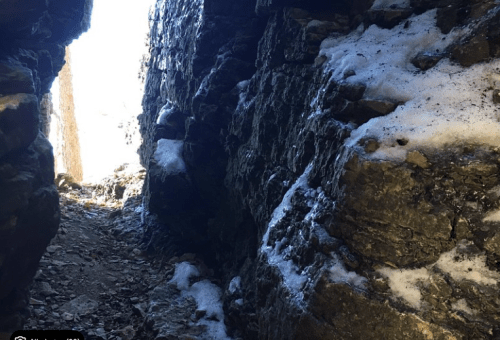 Narrow rocky passageway with snow, leading to a bright light in the distance.