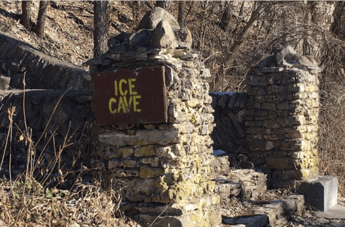 Stone pillars with a sign reading "ICE CAVE" in a wooded area, surrounded by dry grass and trees.