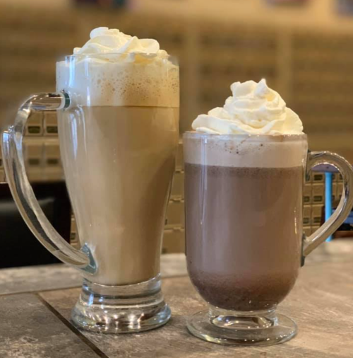 Two tall glasses of beverages topped with whipped cream: one light brown coffee drink and one dark brown hot chocolate.