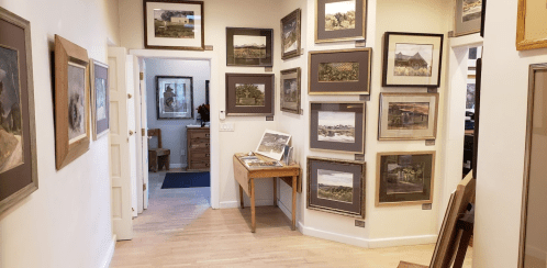 A gallery hallway with framed artwork on the walls and a wooden table displaying more art.