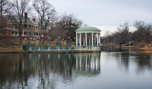 A serene lake scene featuring a gazebo, trees, and a historic building in the background, reflecting on the water.