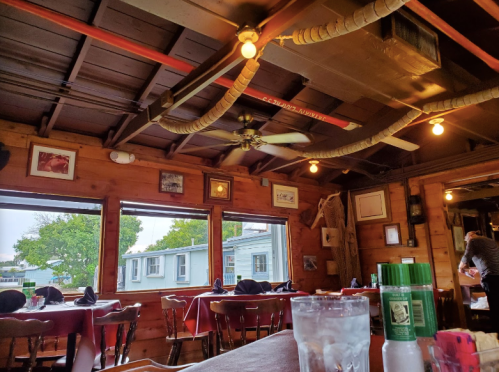 Cozy restaurant interior with wooden walls, ceiling fans, and tables set for dining, featuring large windows and artwork.
