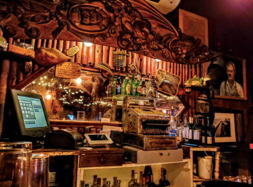 A rustic bar counter with an antique cash register, bottles on shelves, and warm lighting creating a cozy atmosphere.