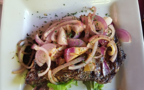 Grilled steak topped with sliced onions and herbs, served on a white plate with a bed of lettuce.