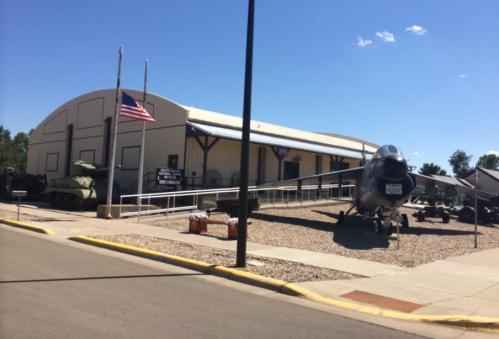 Exterior view of a military museum featuring an aircraft and tanks, with an American flag in front. Clear blue sky above.