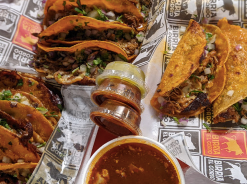A close-up of tacos filled with meat, topped with onions and cilantro, accompanied by sauces and a bowl of broth.