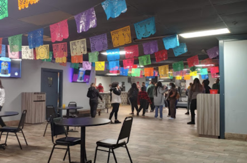 A vibrant indoor space with colorful papel picado decorations, tables, and people socializing.