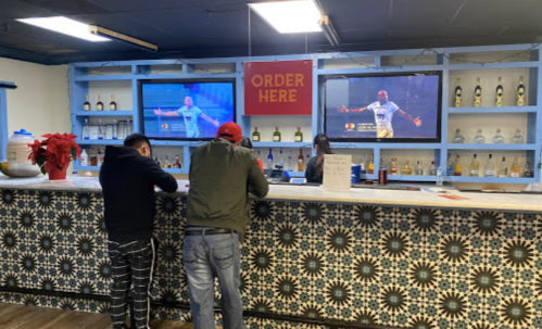 Two people stand at a bar with a patterned counter, watching a sports game on two screens above. A sign reads "ORDER HERE."
