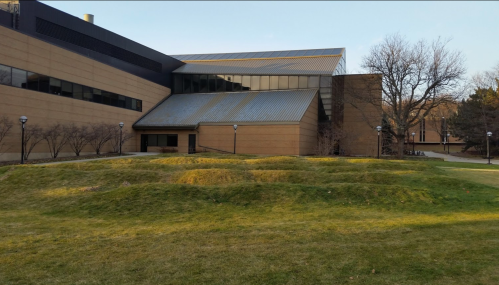 A modern building with a sloped grassy area in the foreground, surrounded by trees and lampposts.