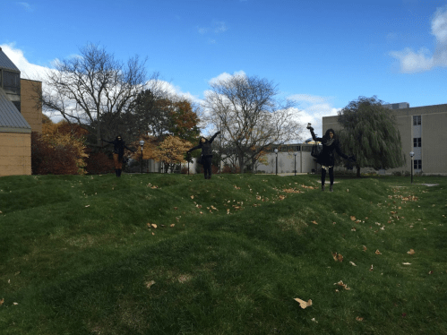 Three people in black clothing playfully pose on a grassy hill under a blue sky with scattered clouds.