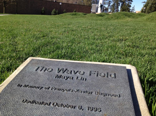 A plaque reading "The Wave Field" by Maya Lin, set on a grassy area, with trees and a building in the background.