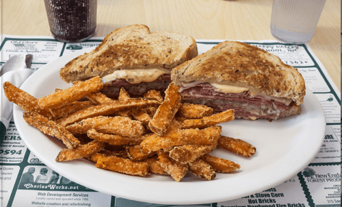 A plate with a toasted sandwich filled with meat and sauce, accompanied by a side of crispy sweet potato fries.