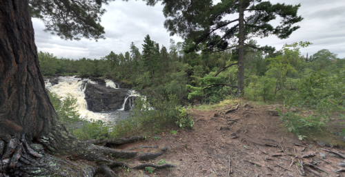 A scenic view of a waterfall surrounded by lush greenery and trees, with a rocky shoreline in the foreground.