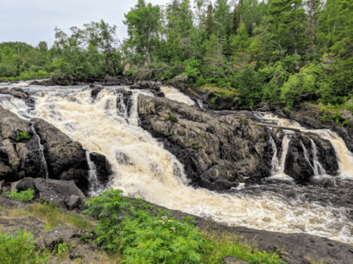 A cascading waterfall flows over rocky terrain, surrounded by lush green trees and vegetation.