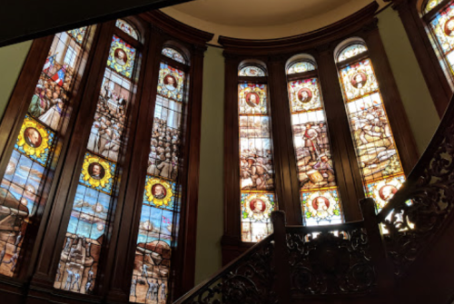 Stained glass windows depicting historical scenes, framed by ornate woodwork and a staircase in a grand interior.
