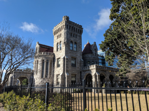Historic stone castle with turrets, surrounded by greenery and a black fence, under a clear blue sky.