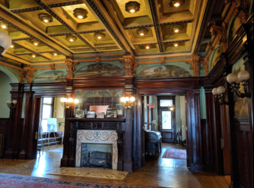 Interior of a historic room featuring ornate woodwork, a decorative ceiling, and a fireplace with artwork on the walls.