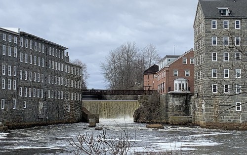 A waterfall flows between historic stone and brick buildings, with a cloudy sky overhead and trees in the background.