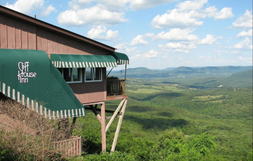 A mountain inn with green awnings overlooks a lush valley and distant hills under a partly cloudy sky.