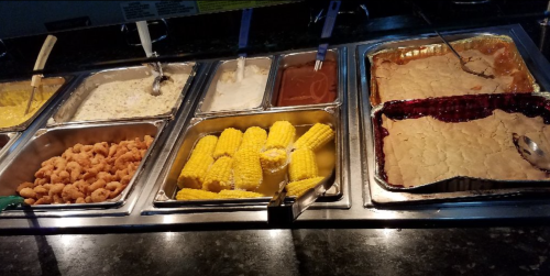 A buffet spread featuring shrimp, corn on the cob, creamy dishes, and dessert in metal trays.