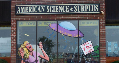 Storefront of American Science & Surplus featuring colorful alien-themed artwork and a sign stating "Open 7 Days a Week."