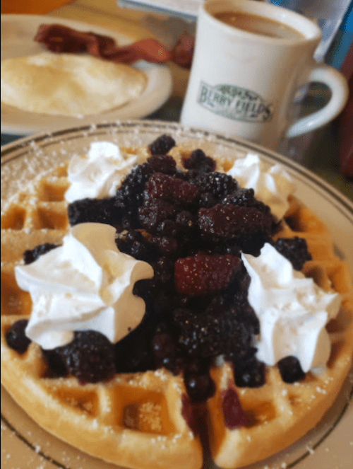 A waffle topped with mixed berries and whipped cream, with a side of bacon and a cup of coffee in the background.
