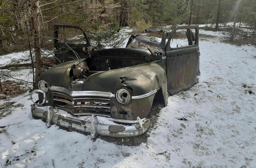 An abandoned, rusted car partially covered in snow, surrounded by trees in a forested area.