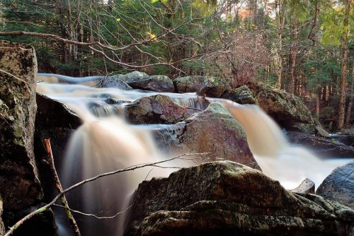 A serene waterfall cascades over smooth rocks, surrounded by lush green trees in a tranquil forest setting.