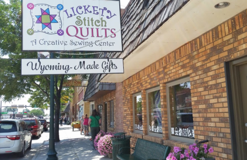 A storefront with a sign for "Lickety Stitch Quilts" and a display of flowers, cars parked along the street.